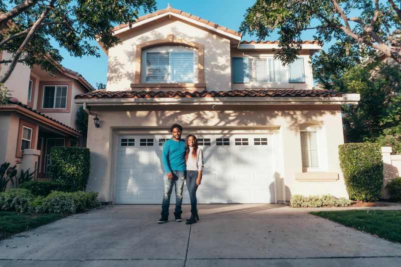 Smiling couple standing outside their new house under a clear blue sky on a sunny day.