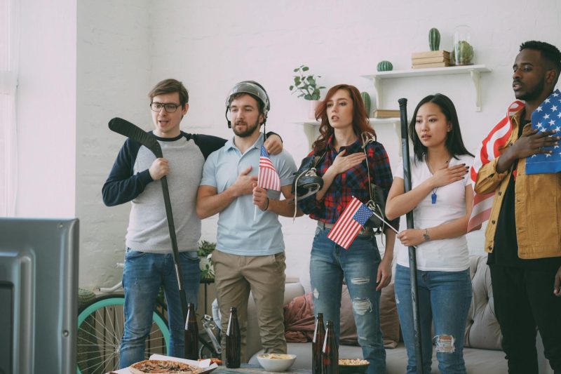 Diverse friends gathered indoors, watching sports on a TV surrounded by American flags.